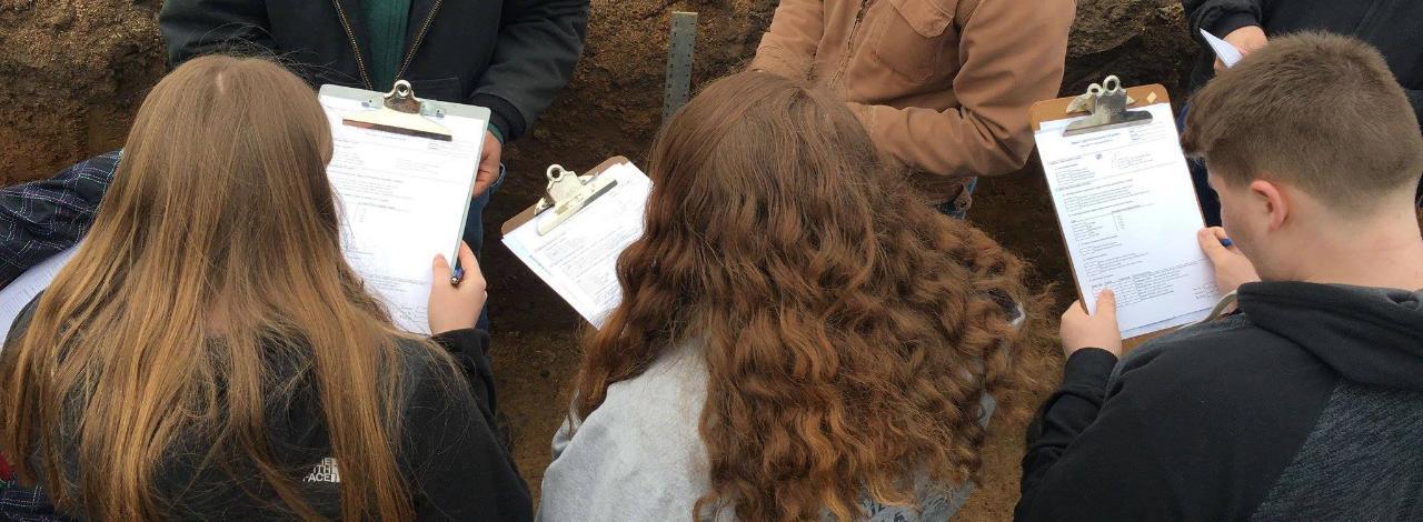 Students taking notes in a soil survey under observation of instructor.