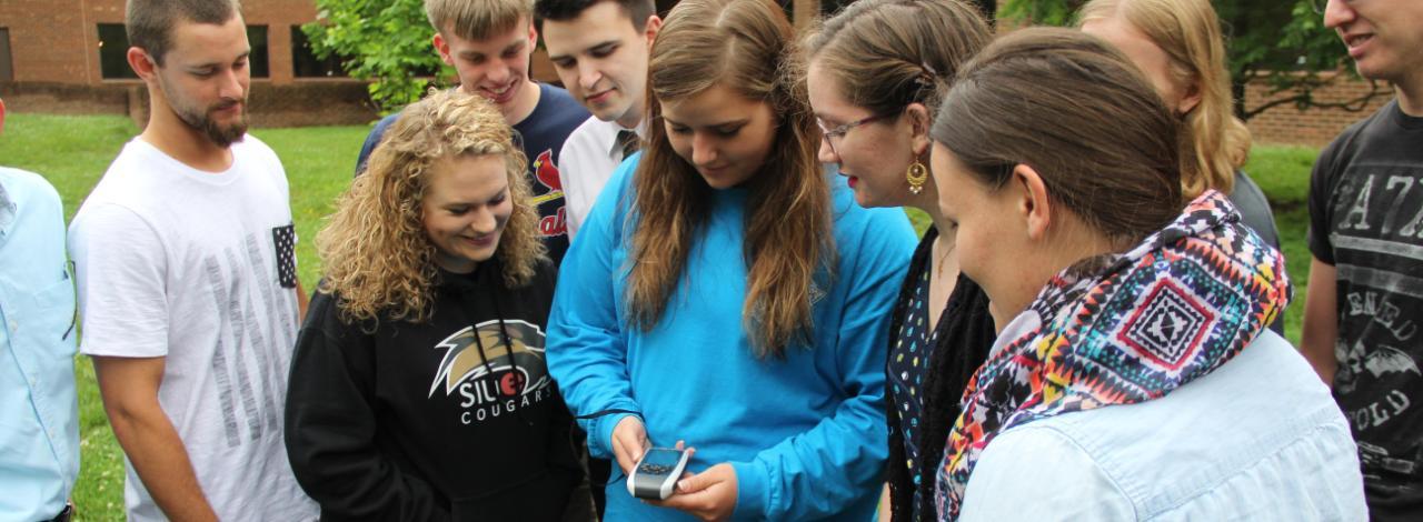 Students gathering around watching student operate handheld device.